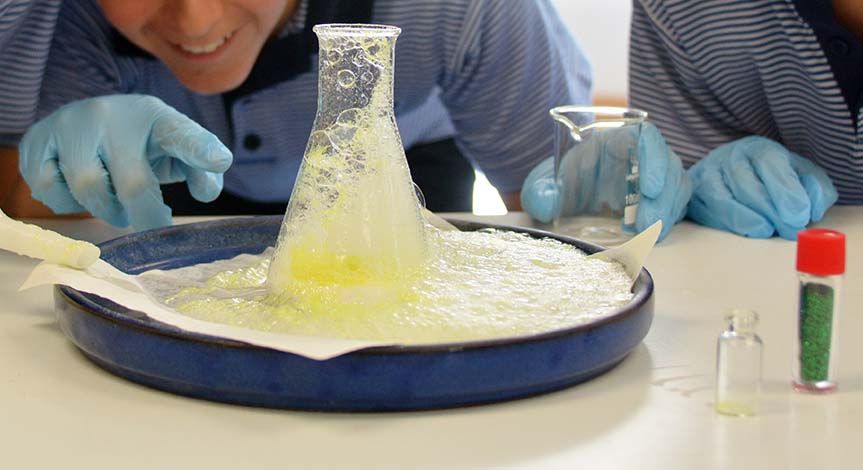 A boy's hand pointing at a beaker with yellow liquid overflowing onto a blue tray
