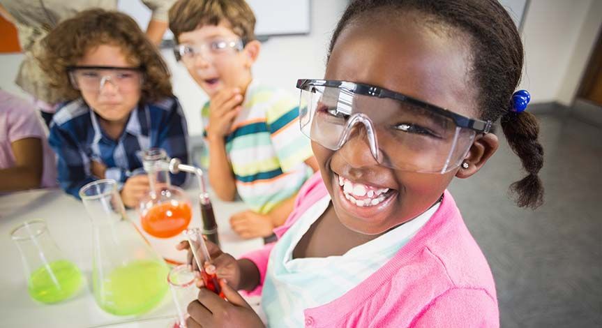 Girl smiling while holding a test tube with liquid inside. 2 other boys looking up at girl.  There are 2 beakers on the desk.