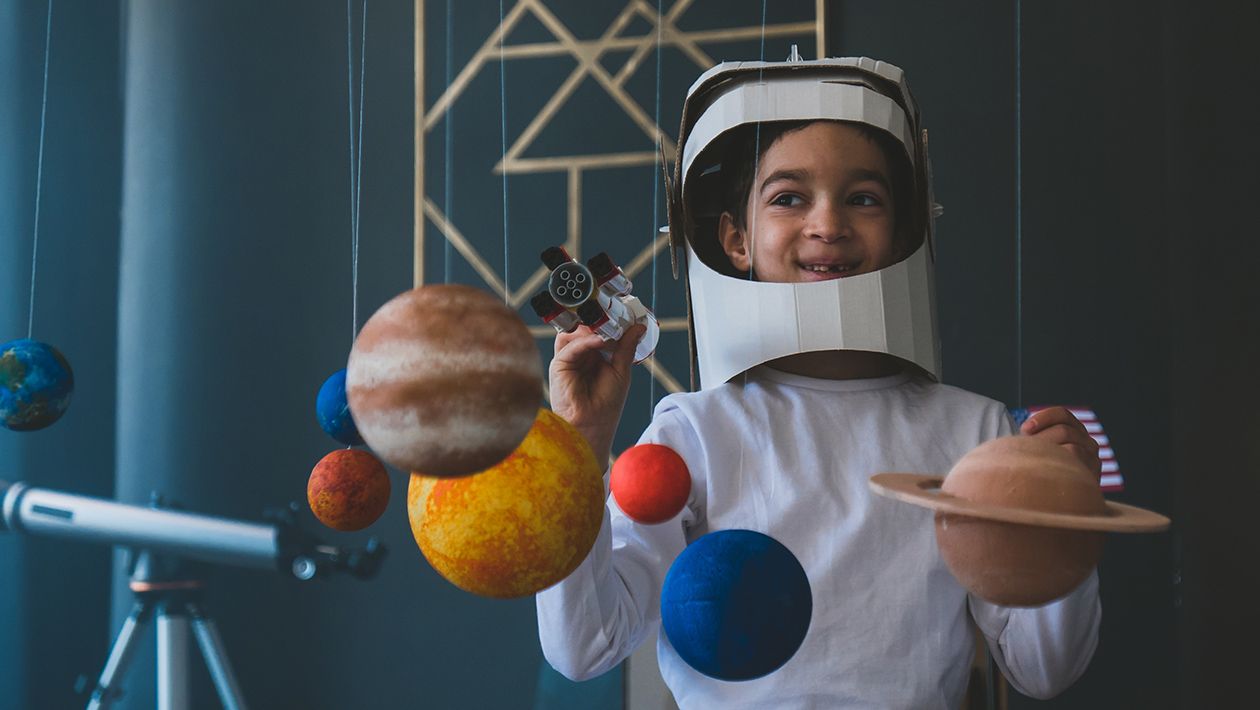 boy wearing cardboard astronaut helmet flying toy rocket through planets, cardboard spaceship rocket in the background
